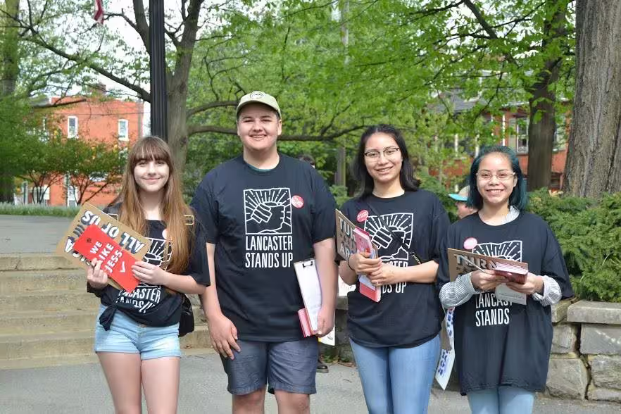 four young people standing in a row, holding clipboards and pamphlets and wearing "lancaster stands up" shirts